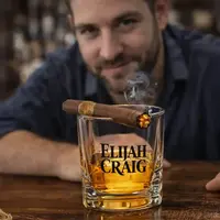 Close-up of whiskey cigar glass with lit cigar resting on top and bourbon inside, man in background, premium bar and distillery promotional product
