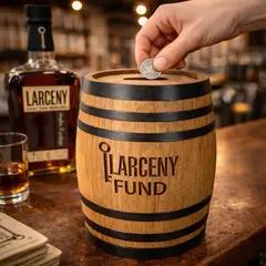 Engraved Larceny Fund oak barrel piggy bank with coin slot on a distillery bar as a hand drops a coin in, showcasing branded distillery gift shop merchandise