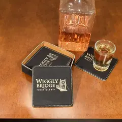 Personalized leather coaster set with whiskey glass and decanter on a wooden table, ideal for custom barware, corporate gifts, and distillery merchandise