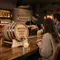 Branded whiskey barrel promotional product in a bar setting pouring bourbon into a glass, with bartender and guests creating a premium brand experience.